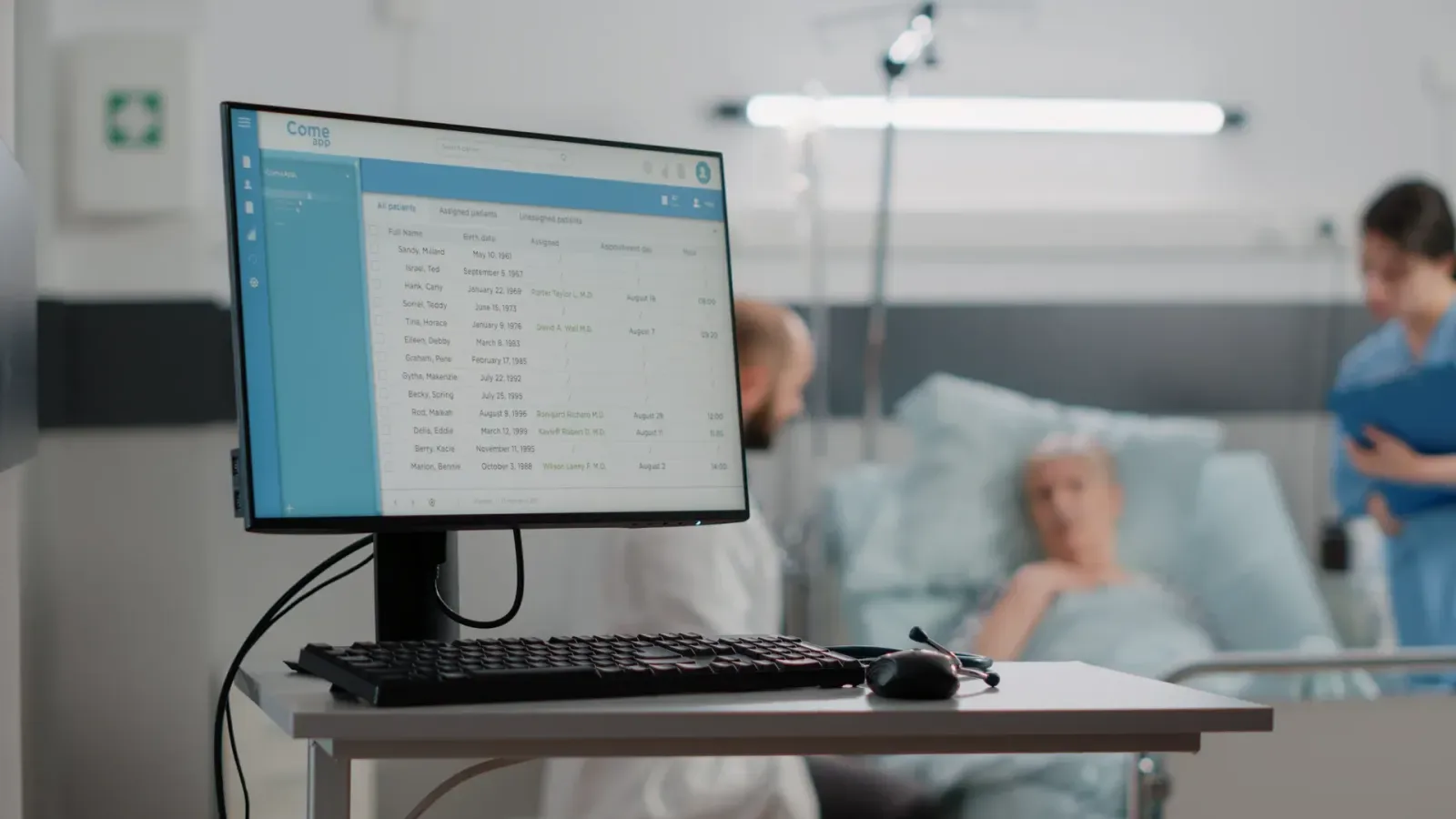 A clinical workstation showing a patient record on screen beside a hospital bed, with care staff in the background