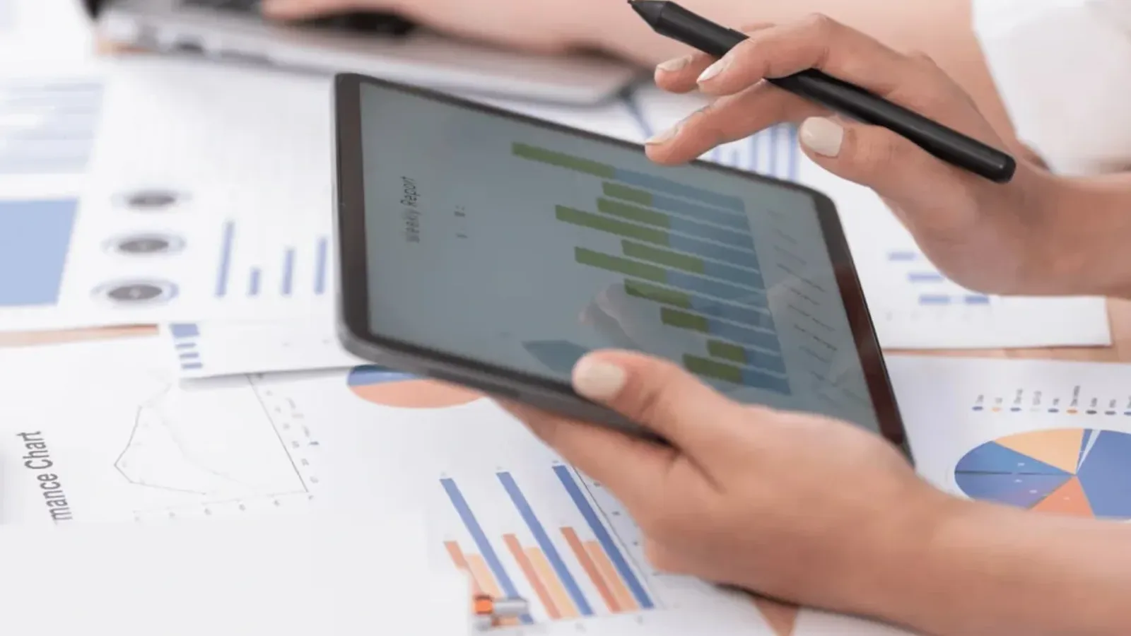 Hands holding a tablet showing bar charts above printed financial reports on a desk