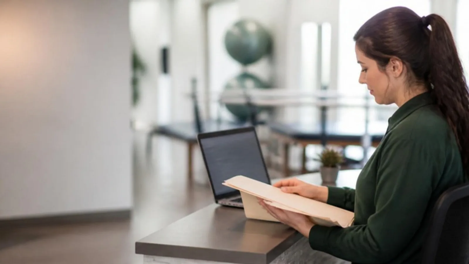 Front-desk coordinator at a physical therapy clinic organising a paper case file beside an open laptop, with therapy equipment softly blurred behind