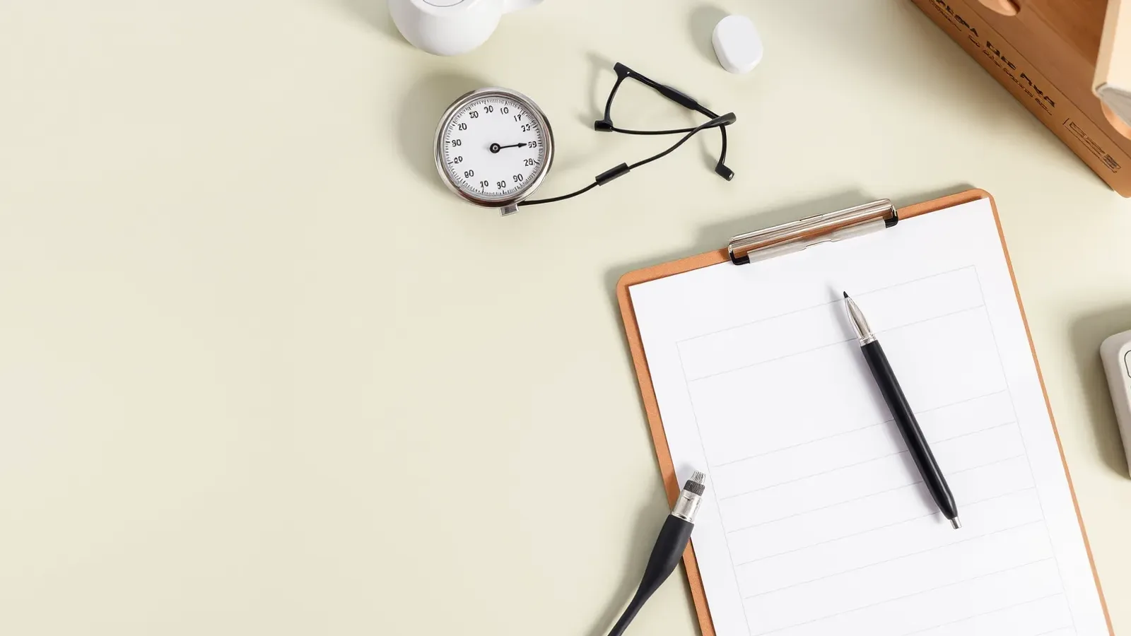 Overhead view of a clipboard with a blank form, a pen, reading glasses and a blood pressure gauge on a cream desk