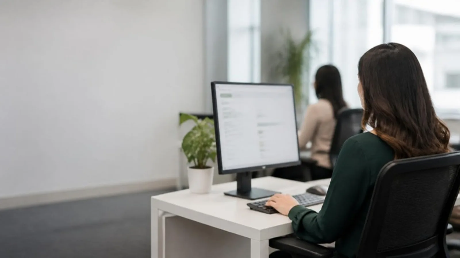 Claims examiner at a clean health plan office desk reviewing a claims queue on a monitor, with a colleague at the next desk in soft focus