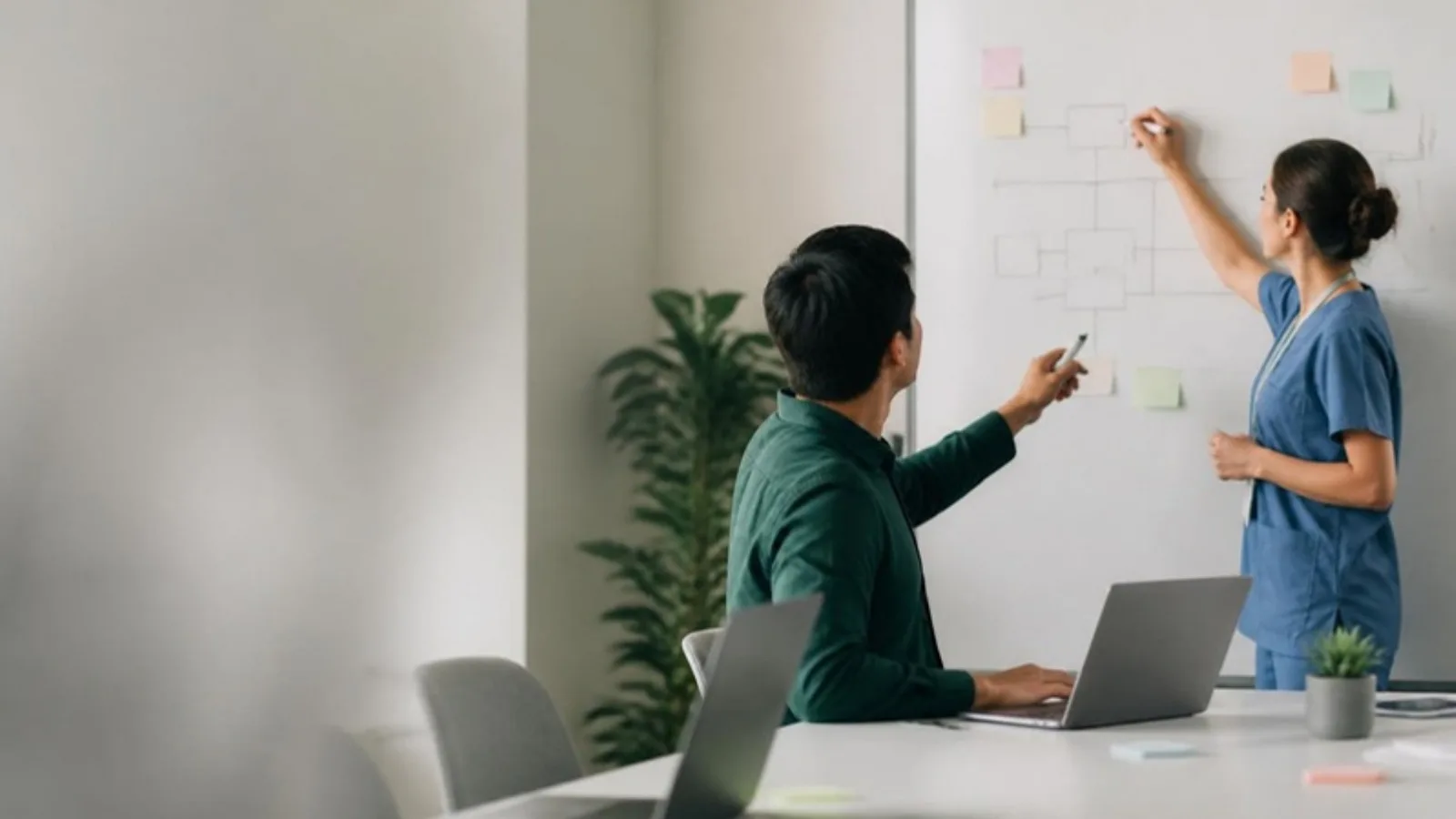 Clinician in scrubs and a developer sketching a care-pathway prototype on a whiteboard with sticky notes, laptops open on the table
