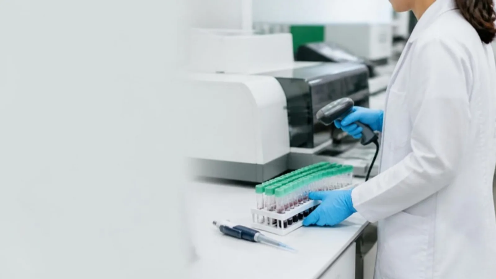 Laboratory technician in gloves scanning a barcoded rack of sample tubes with a handheld scanner beside benchtop instruments
