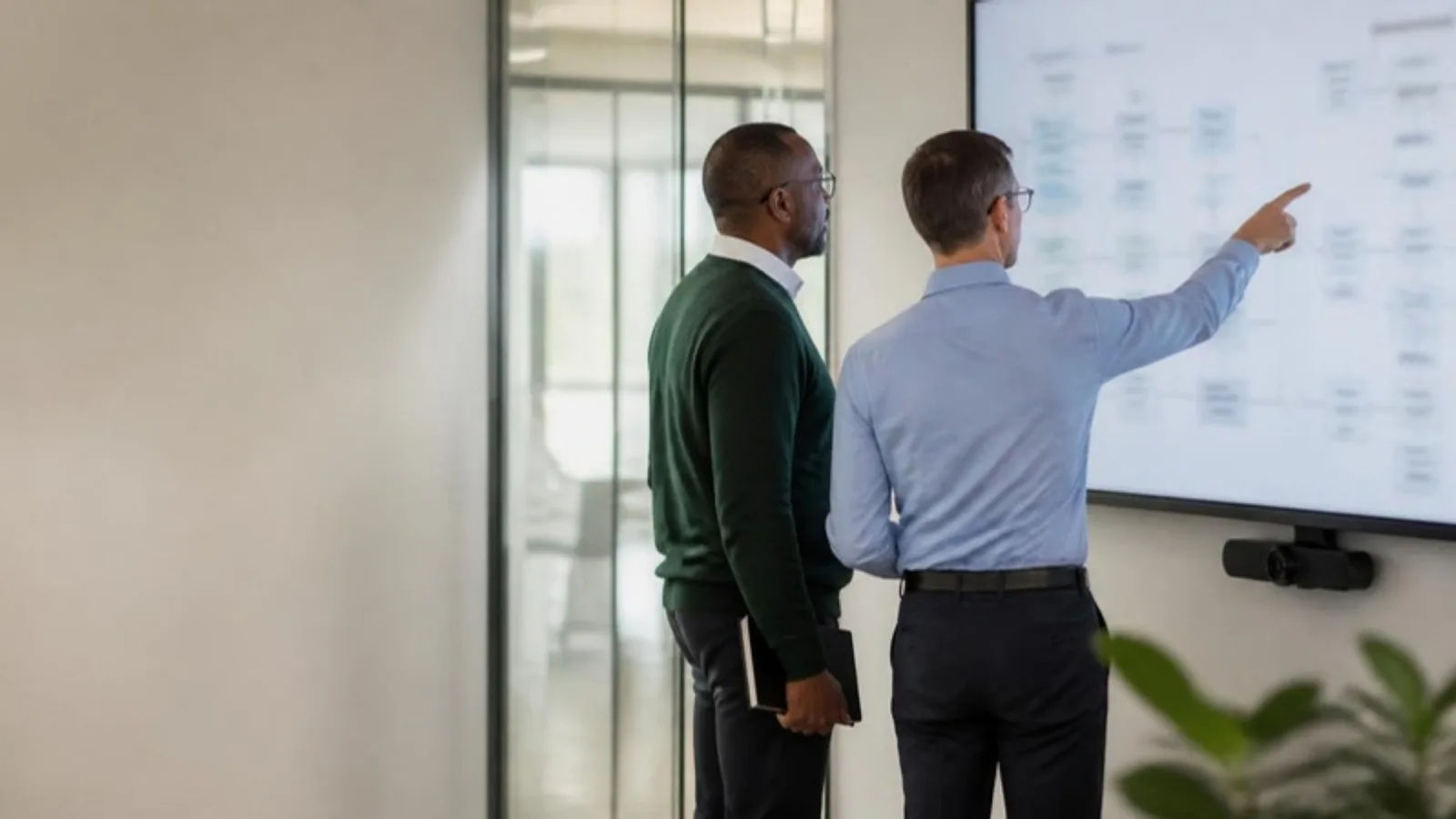 Two health plan technology leads in a glass meeting room reviewing a system architecture diagram on a large wall display