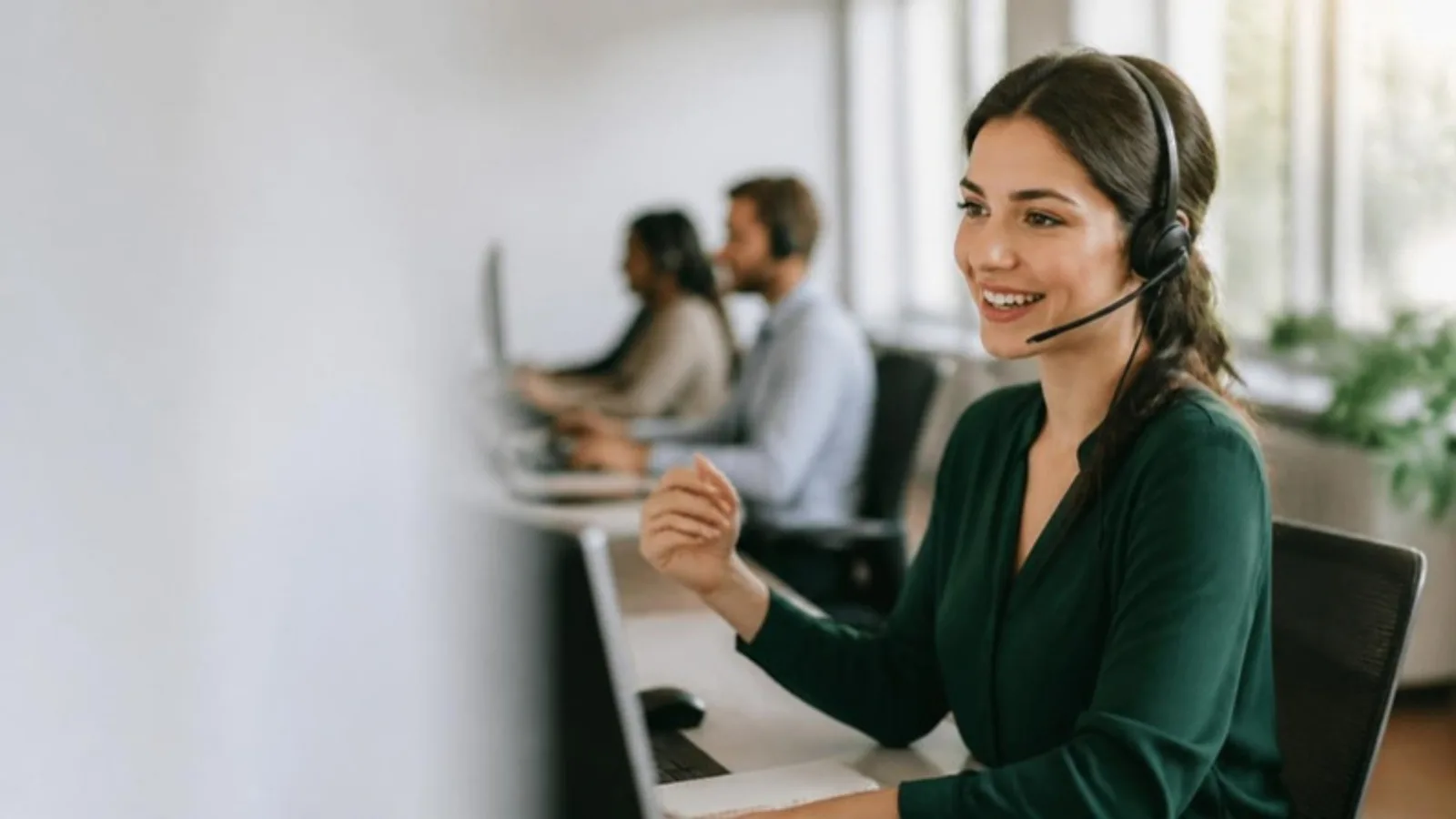 Member services agent wearing a headset at a bright call-centre desk, with other agents at their desks in soft focus behind her