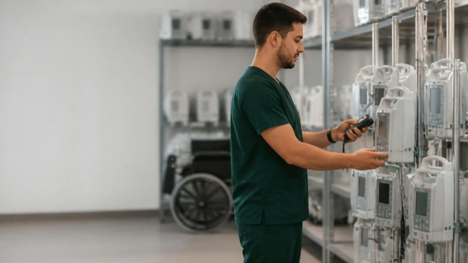 Biomedical technician in green scrubs scanning an RFID tag on an infusion pump with a handheld reader in a hospital equipment room