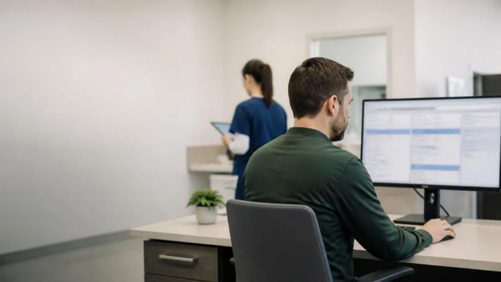Salesforce administrator at a hospital office desk configuring a CRM application on a widescreen monitor, with a clinical colleague in scrubs in soft focus behind