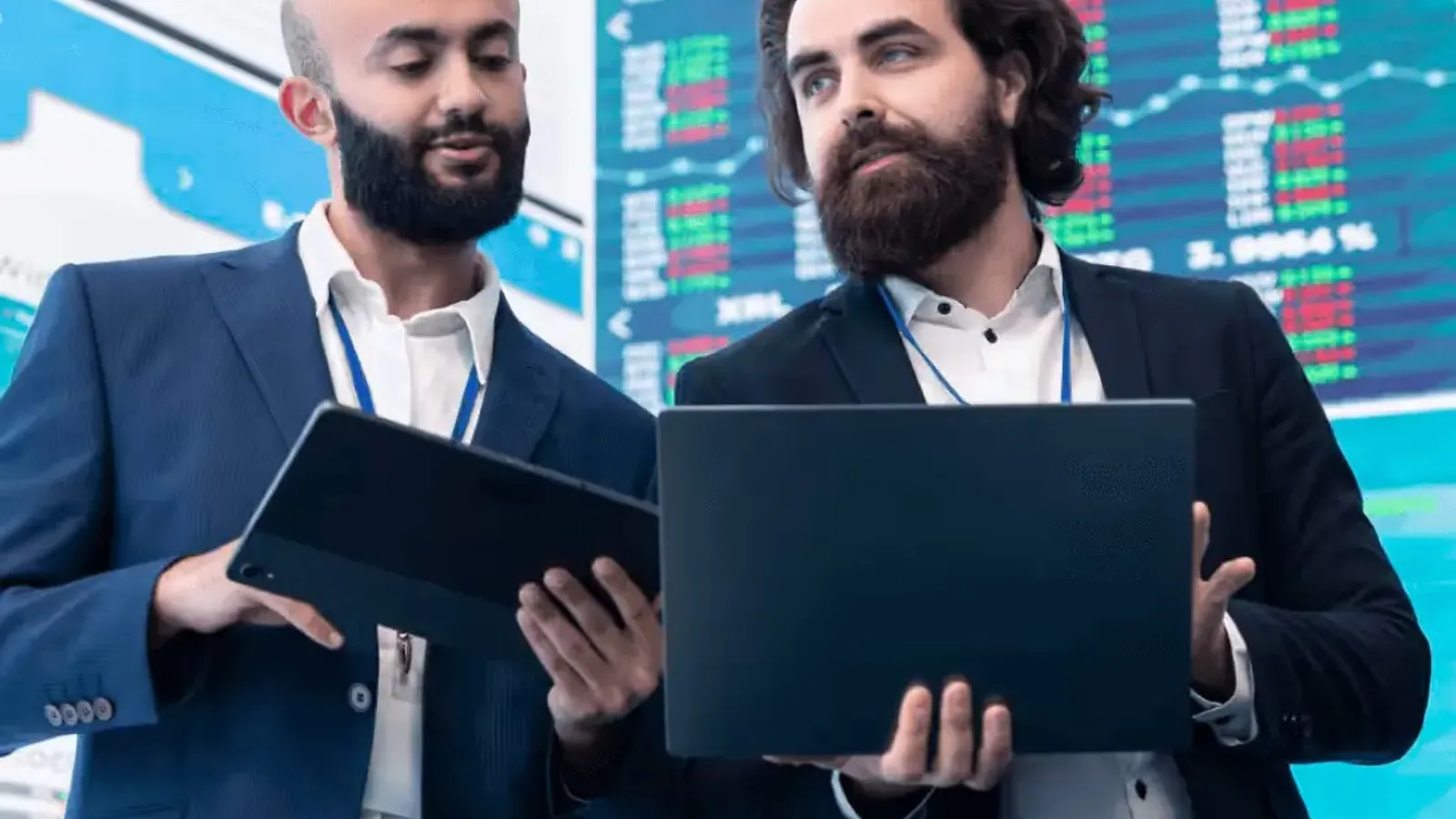 Two business professionals reviewing financial data on a tablet and a laptop in front of market screens