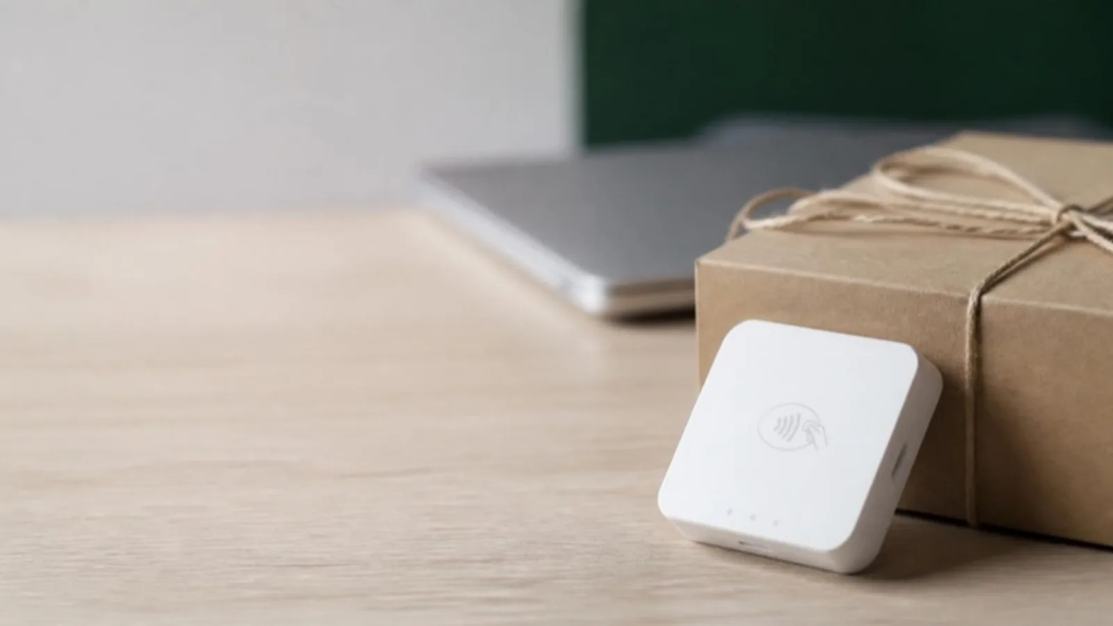 Close-up of a contactless card reader beside a sealed kraft shipping box on a pale wooden packing bench, with a closed laptop behind