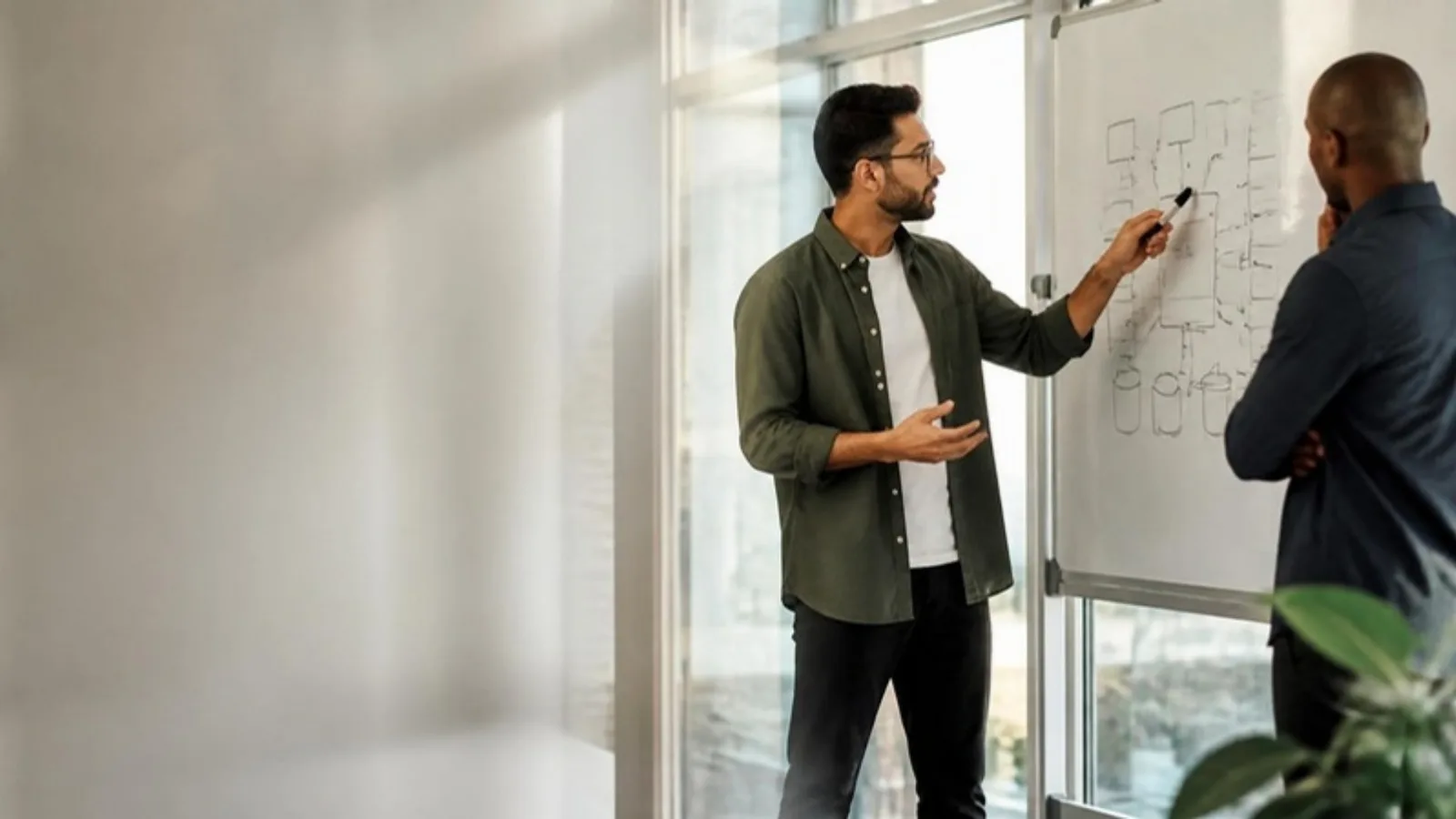 Cloud architect explaining an architecture diagram on a whiteboard to a client in a glass-walled office