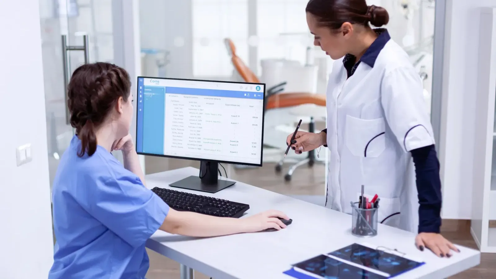 Two clinic staff members at a front desk computer showing a patient schedule